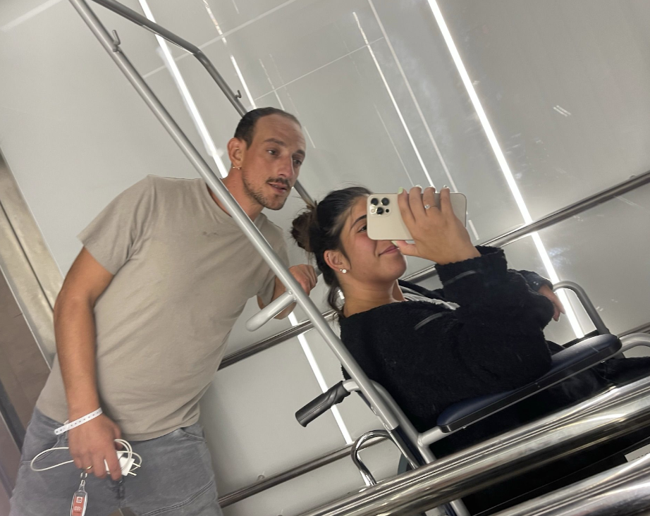 Danielle is sitting on a chair with her husband standing next to her